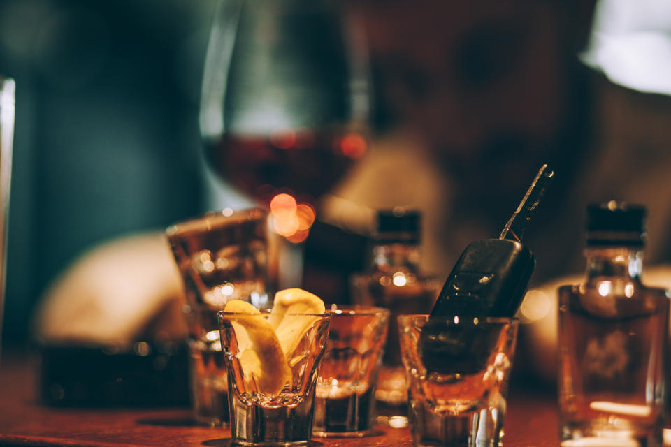 A dimly lit bar scene with a blurred person in the background, liquor bottles, a wine glass, and shot glasses containing lemon slices and a phone