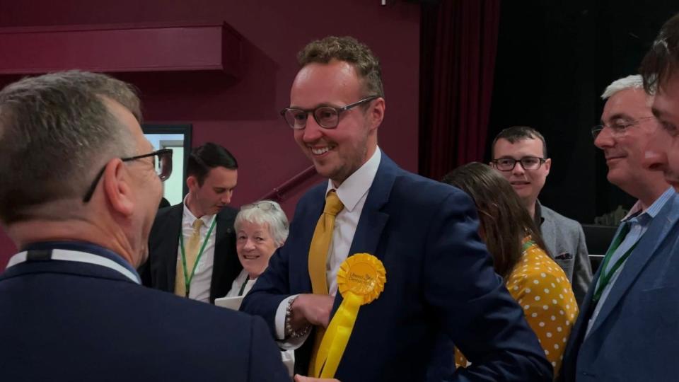 Image of Adam Dance. He has light hair, facial hair and glasses. He is wearing a suit, yellow tie and a yellow Liberal Democrat badge. 