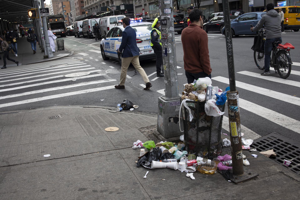 A city street with litter on the ground near a trash can. A police officer and a person crossing the street. Police cars and bicycles in the background