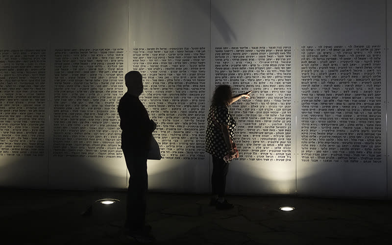 In a photo taken at night, a wall is seen with many names listed on each panel. spotlights on the ground shine upward to illuminate the names. Two people stand in front of the wall, and one of them is reach forward to point out a name.