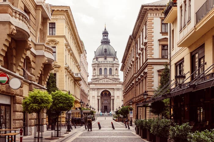 View of a European street leading towards a large dome-capped building, flanked by classic architecture and a café-lined sidewalk