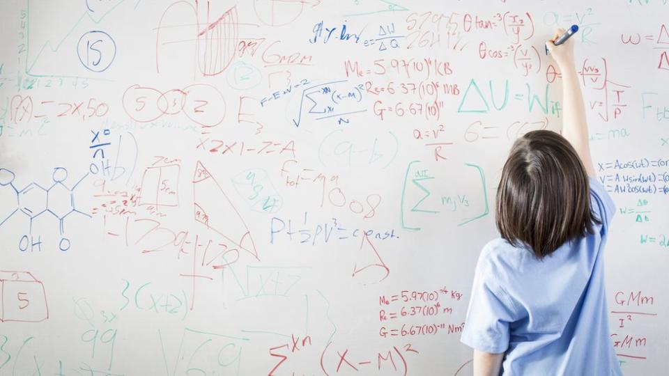 Un pizarrón enorme blanco lleno de fórmulas matemáticas y una niña escribiendo sobre él.