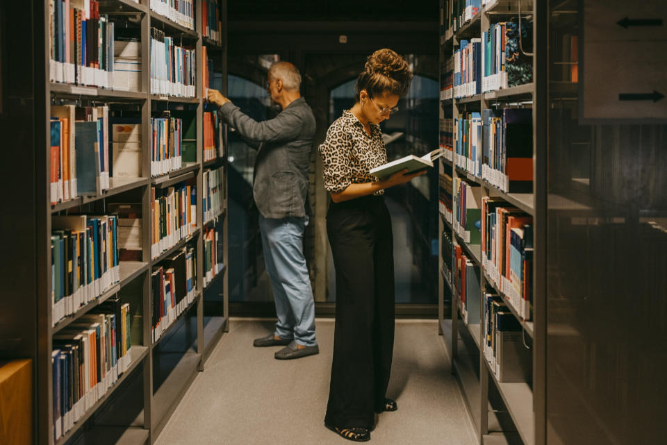 Two people are in a library. A woman reads a book while standing. A man searches the shelves in the background. Their names are not provided