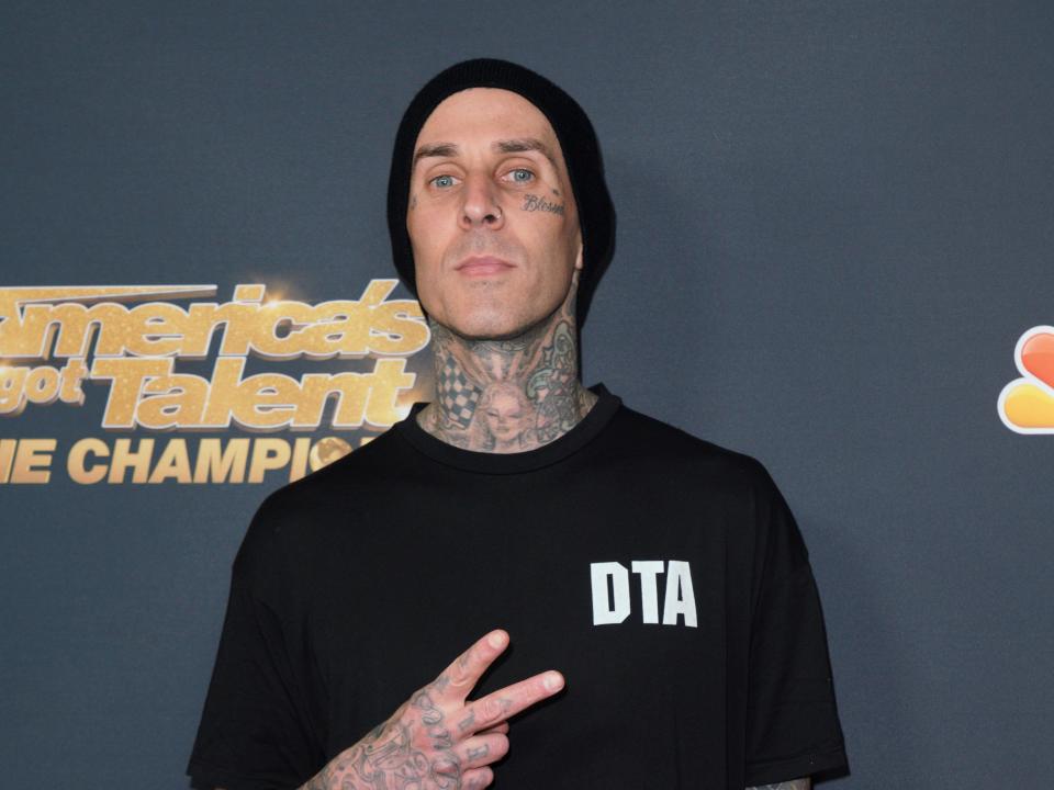 travis barker on the america's got talent red carpet. he has tattoos on his arms, neck, and face, and is flashing a peace sign with a neutral expression on his face. he is wearing a black beanie, as well as black pants and a black shirt that says "DTA" on the left breast