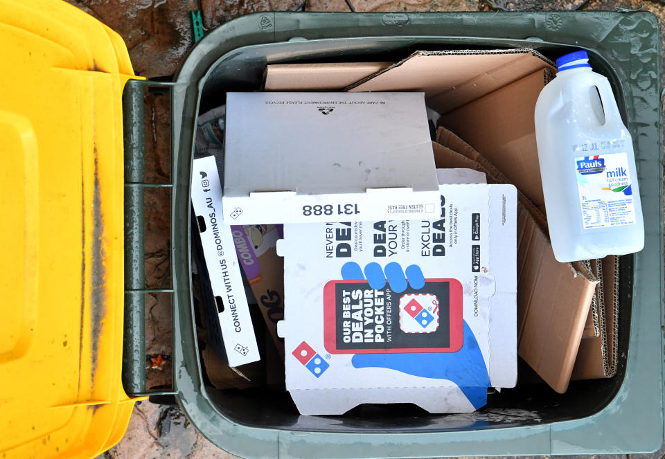 A yellow recycling bin can be seen with cardboard and plastic milk bottles in it. 