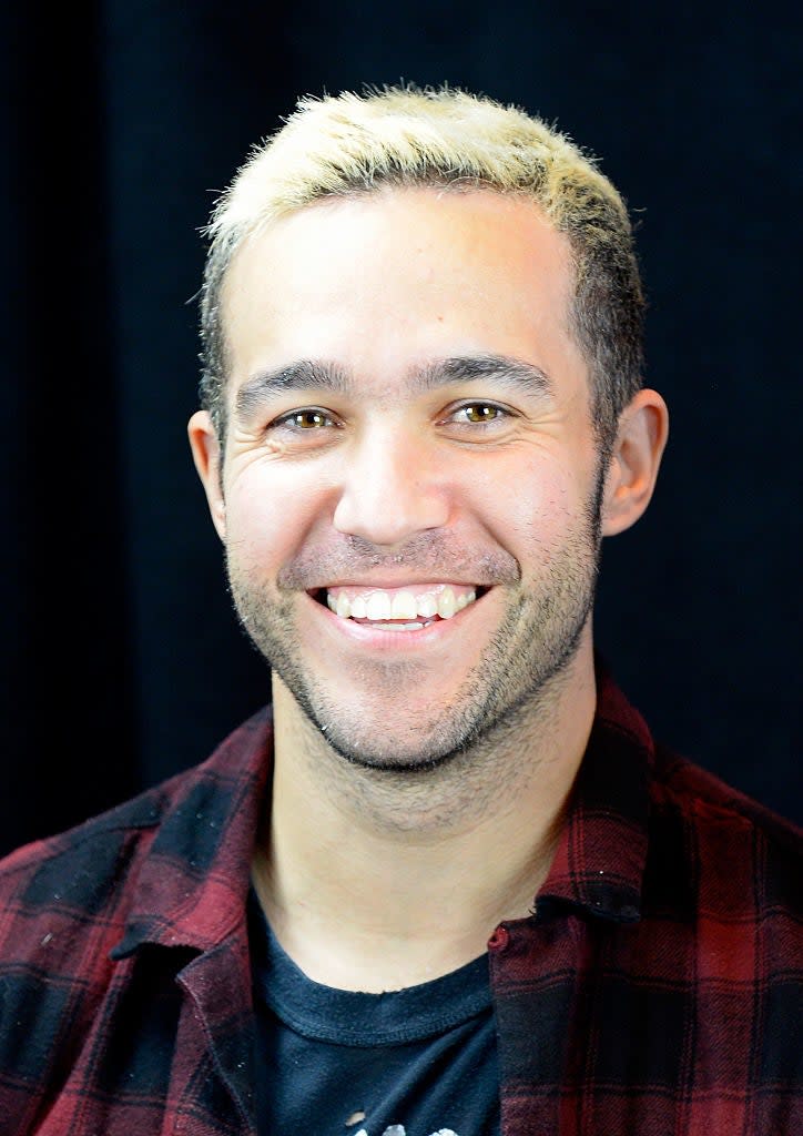 Pete Wentz smiling, wearing a flannel shirt over a graphic T-shirt, against a dark background