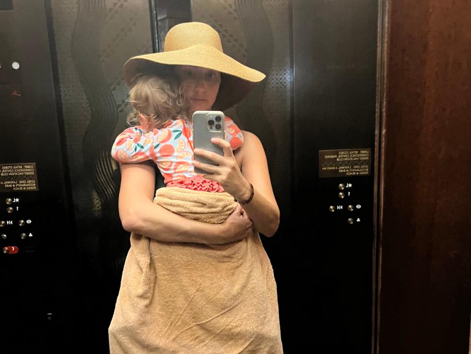 A woman wearing a big floppy hat taking an elevator selfie with her sleeping child.