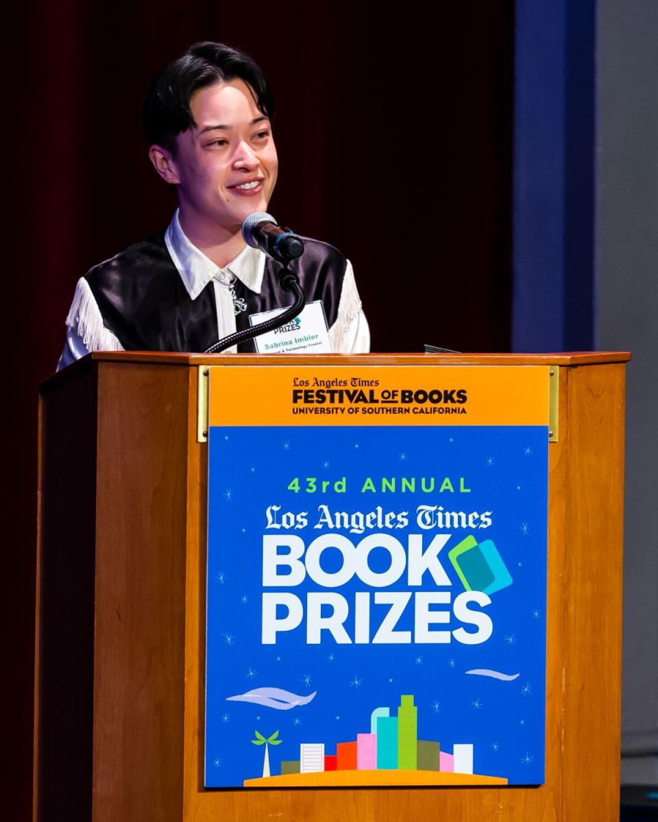 A woman in a tuxedo-print shirt stands behind the podium.