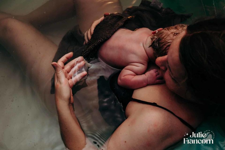 Mother cradling newborn on her chest in a birthing pool, with a caregiver's hand supporting