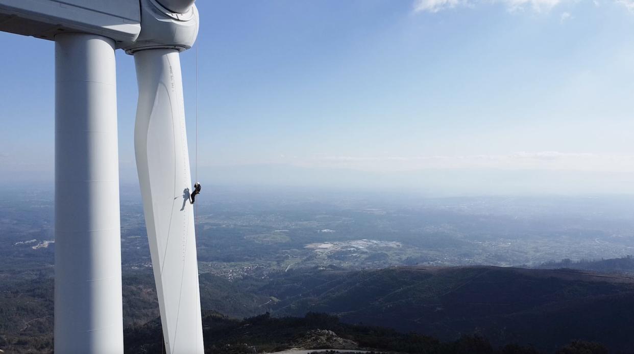 A wide shot of a lone wind turbine technician hanging on the side of a wind turbine in the mountains of Portugal.