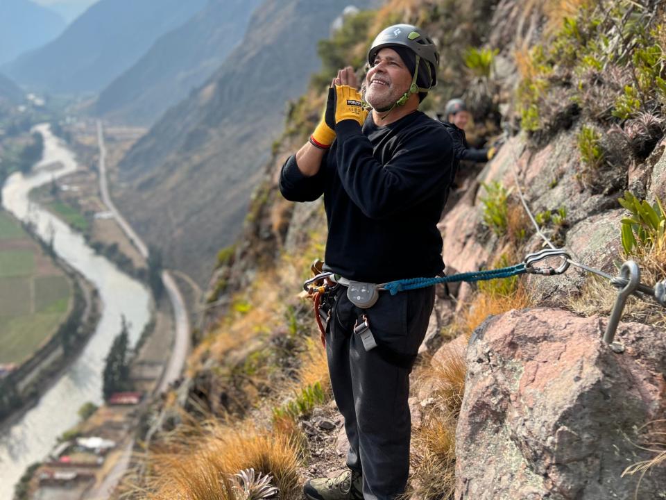 A man holding up hands to pray while climbing via ferrata on the side of a cliff.