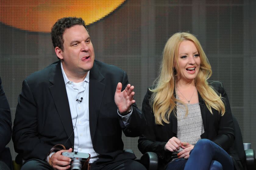 A man with short brown hair in a suit gesturing with his hand and sitting next to a woman with long blond hair in a blazer