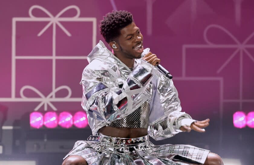 A man in a silver jacket and skirt singing into a microphone on a stage