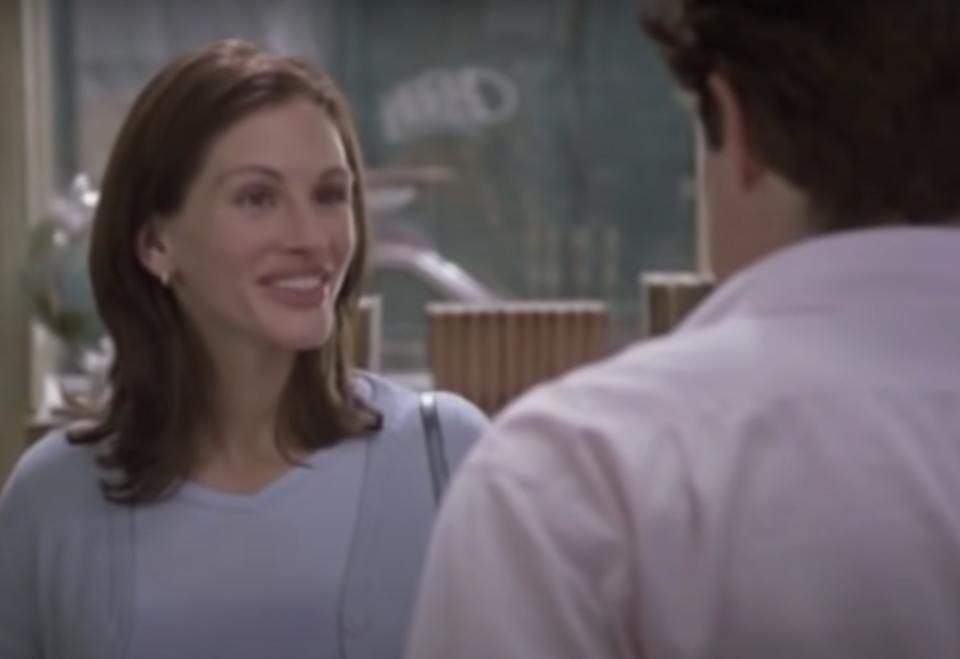 Actor Julia Roberts wears a blue top and sweater and smiles at Hugh Grant whose back is facing the screen.