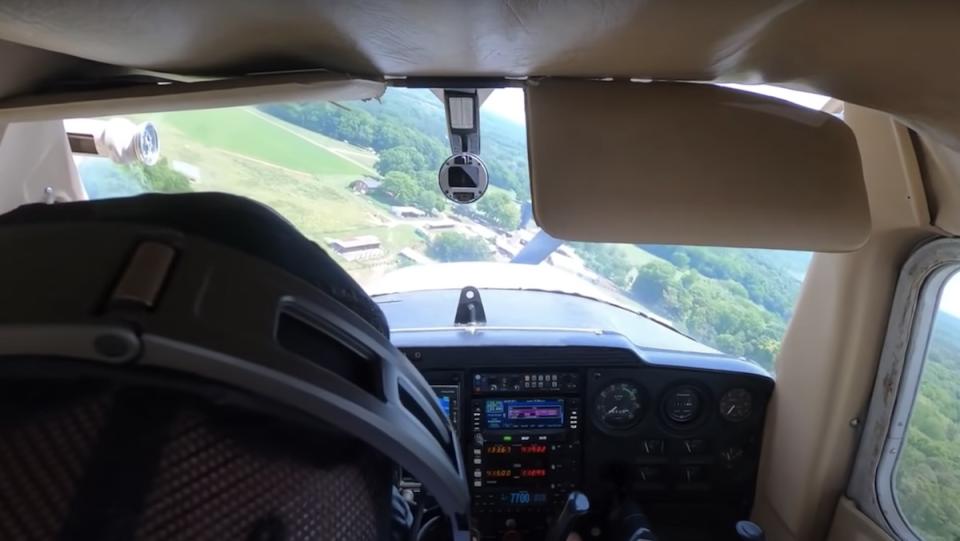 Behind the head of a pilot inside a small plane looking out over green fields
