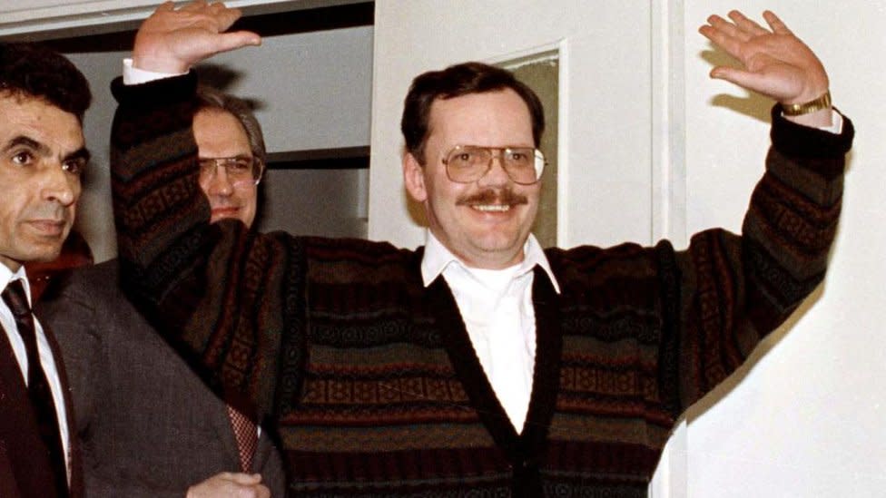 A man in glasses holds his hands up as he walks through a door