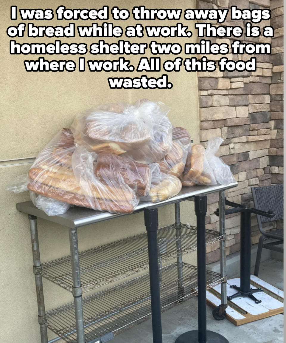 A metal cart outside with several loaves of bread wrapped in plastic bags piled on top, next to a brick wall and a chair