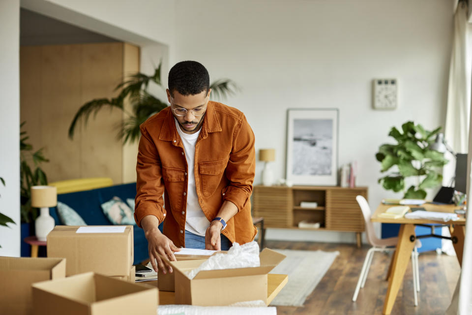 Young male entrepreneur packing cardboard box at table in apartment. 