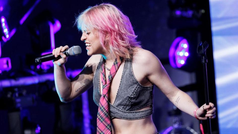 Beth McCarthy on stage.  Beth is a 26-year-old white woman with blonde hair dyed pink.  She wears a gray denim crop top with a pink tartan tie around her neck.  She holds a microphone in front of her face with her right hand, revealing a black line tattooed on the inside of her arm.  Beth's eyes are closed as she sings, holding the microphone stand with her left hand.  The scene behind her is lit purple