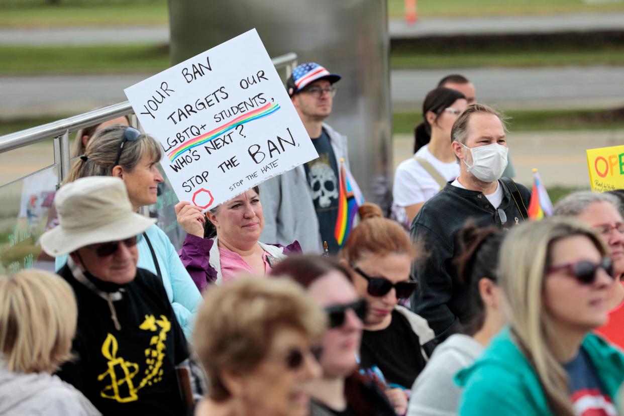 A dozen or so people hold rainbow flags, and signs, one of which reads: Your ban targets our LGBTQ+ students, who's next? Stop the ban. Stop the hate. 