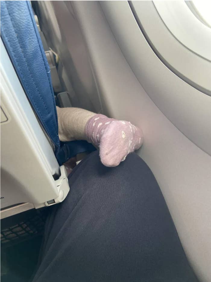 A toddler's socked foot and leg protruding from the side of an airplane seat and  almost resting on an adult's knee