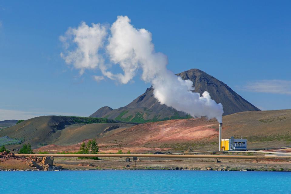 lots of steam arises from a geothermal power station located along water. There's a mountain in the background