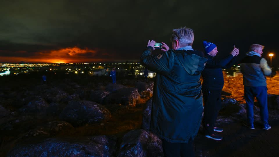 People on the outskirts of Reykjavik take pictures of the orange colored sky as molten lava flows out from a fissure on the Reykjanes peninsula north of the evacuated town of Grindavik, western Iceland, on March 16. - Halldor Kolbeins/AFP/Getty Images