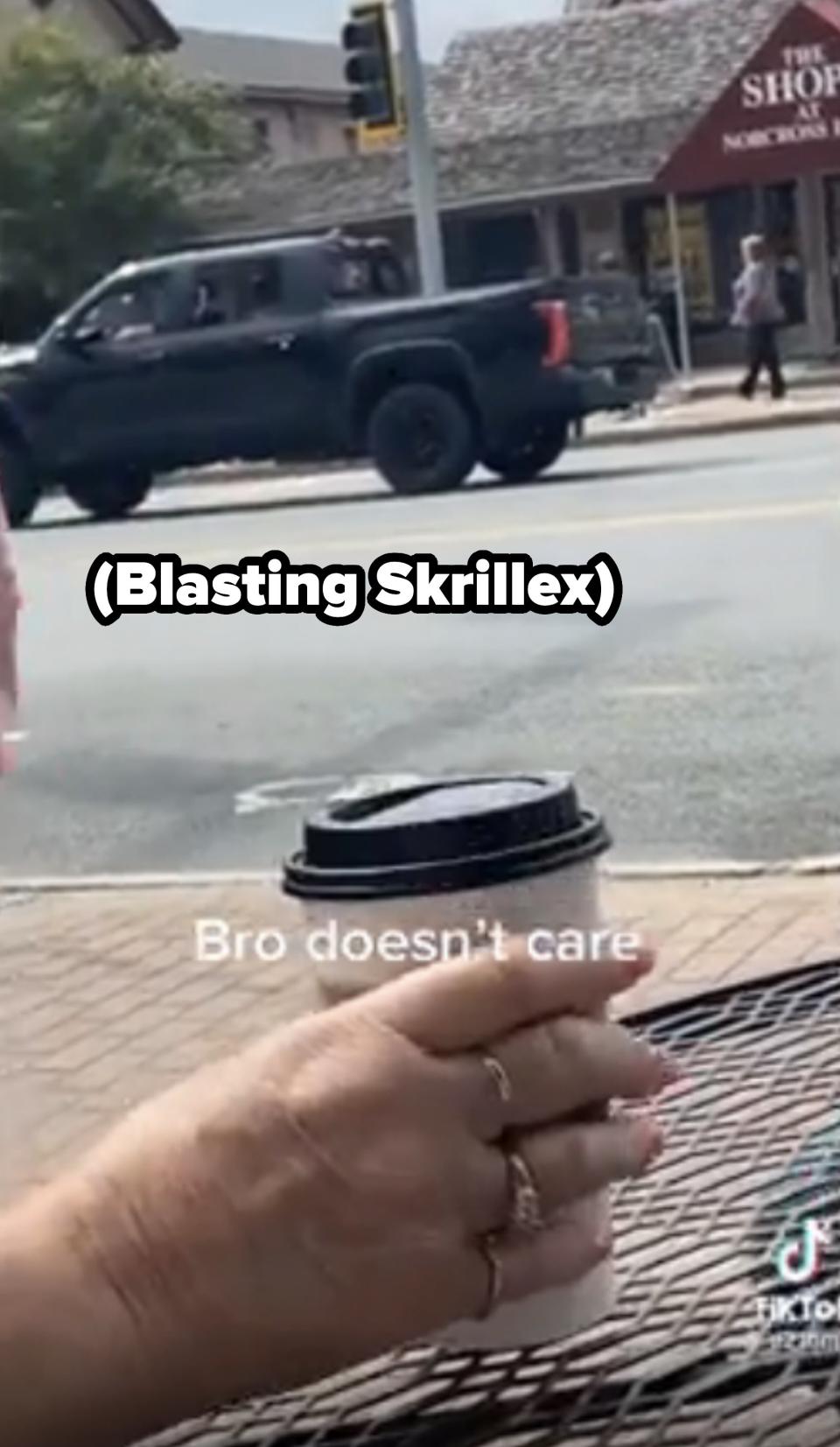 Close-up of a hand holding a coffee cup at an outdoor table with a street in the background. Overlay text reads: "Bro doesn't care."