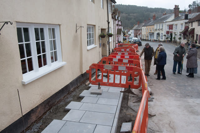 FILE PICTURE - Stone slabs replacing the cobbled pathway in front of houses and businesses in West Street, Dunster.  Paving slabs which replaced iconic cobbles in an historic medieval village are to be removed by a cash-strapped council at a cost of ï¿½45,000 - because they are "too blue".  See SWNS story SWPAVING.  The original stones which dated back to the Bronze Age was torn from a main road in Dunster, Somerset, to improve pedestrian safety and disabled access.  The stones were replaced with blue-hued slabs, which locals complained were too blue, angular, regular, smooth and "like a path on a new housing estate".  Villagers accused the council of 'vandalism' and Historic England was furious that it was not consulted before the work began.  Now, just ten months later, the council has agreed to change the paving slabs to stones which are more in-keeping with the village - at a cost to taxpayers of ï¿½45,000.  It comes after the cash-strapped Tory approved budget cuts of ï¿½16 million earlier this year. 