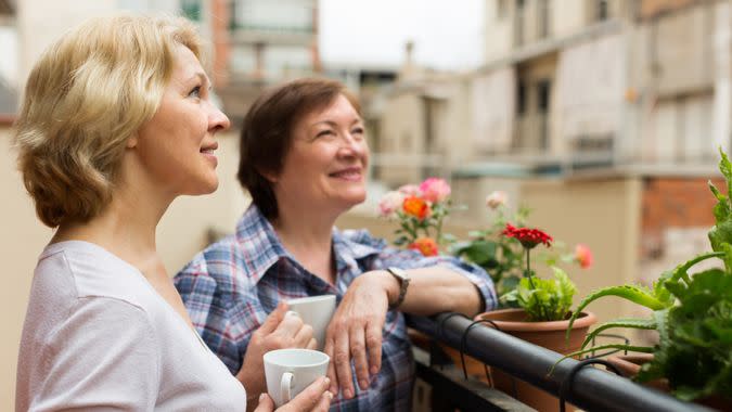 Smiling aged female friends relaxing with cup of coffee on balcony.