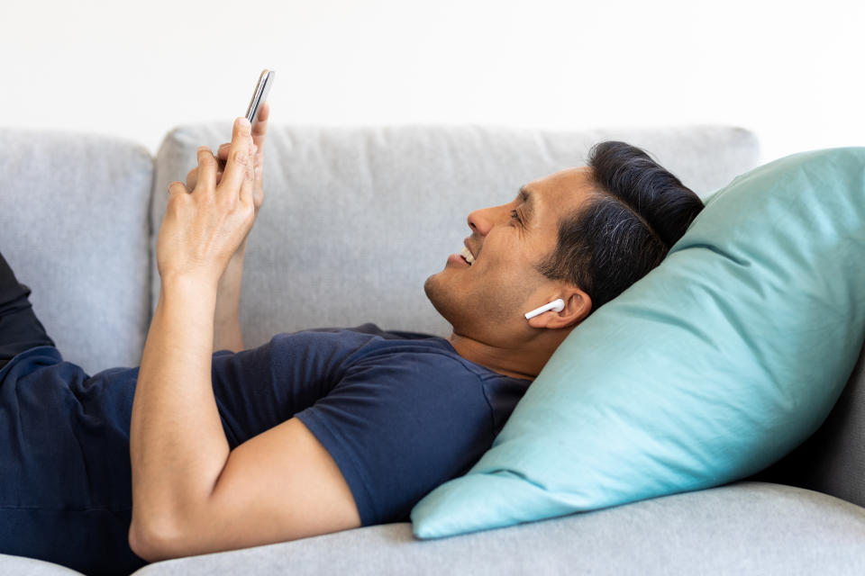 Man lying on sofa using mobile phone at home during pandemic lockdown. Man resting on couch holding cellphone and listens music through wireless earphones.