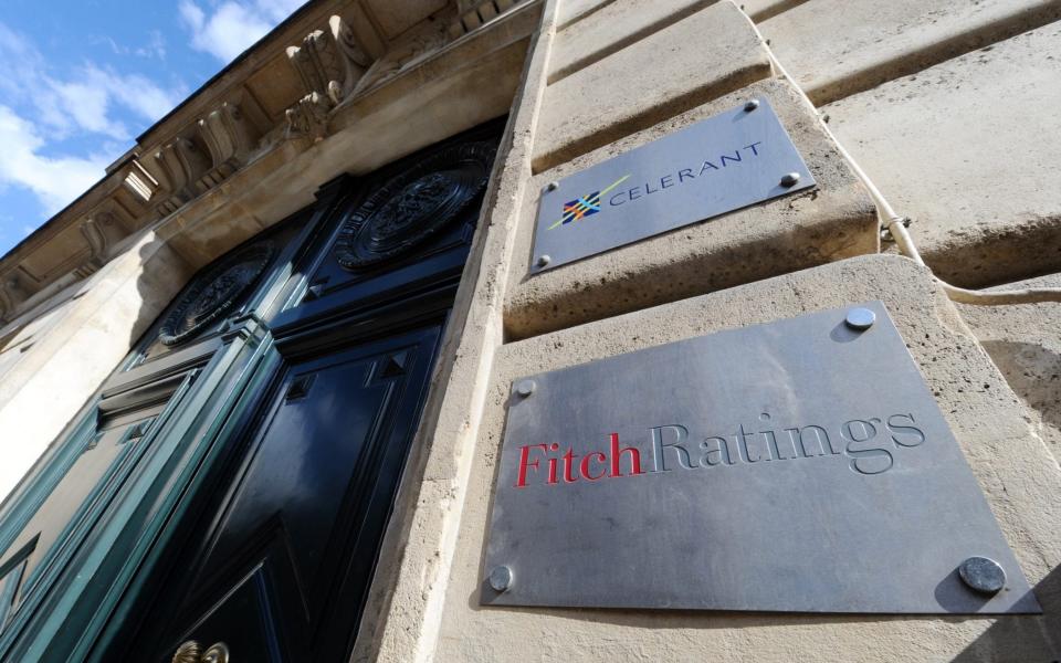 A grey plaque with "Fitch Ratings" engraved on it is bolted to a stone wall next to a towering and ornate black door, part of an old-fashioned building above which a small wedge of bright sky can be seen - Miguel Medina/AFP/Getty