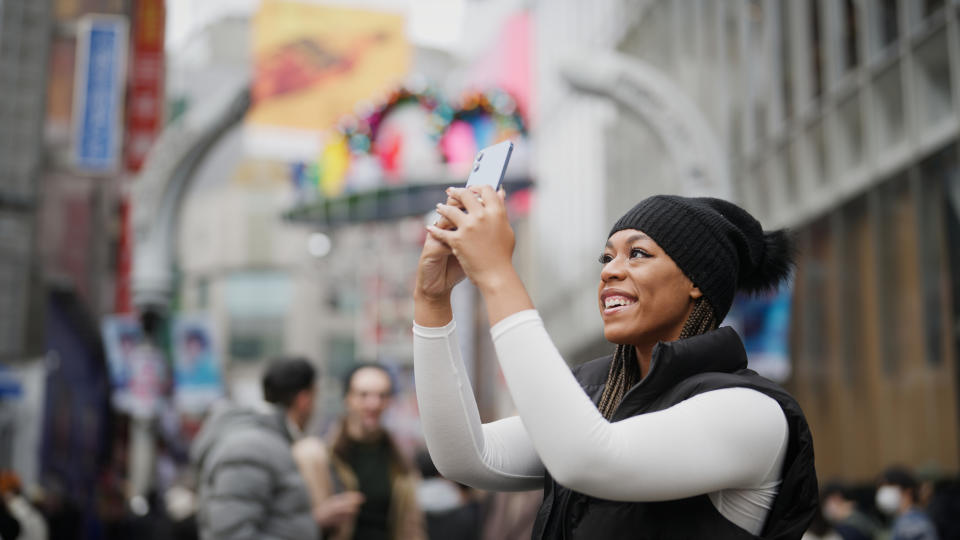 Person holding phone and smiling while taking a photo in a busy city street with blurred people and colorful decorations in the background