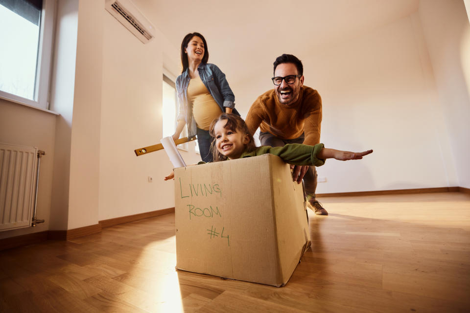 Happy expecting family having fun after moving into a new home. Father is pushing their daughter in cardboard box. Copy space.