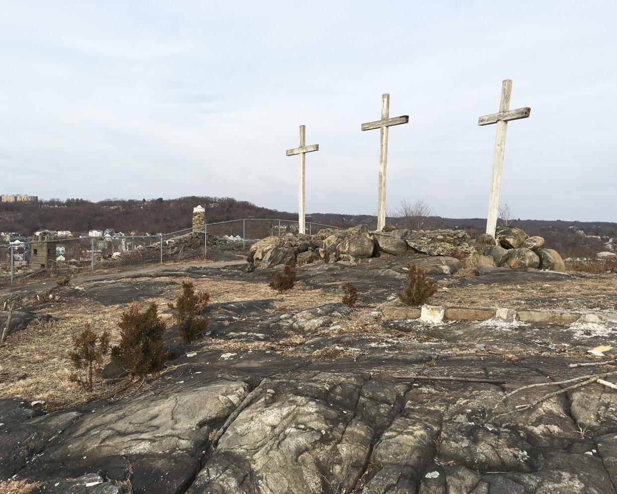 Abandoned Holy Land USA, Waterbury, Connecticut, three large white wooden crosses on rocks, rocky terrain in the foreground, town, trees on hills, and a clouds in a blue sky in the background, during winter