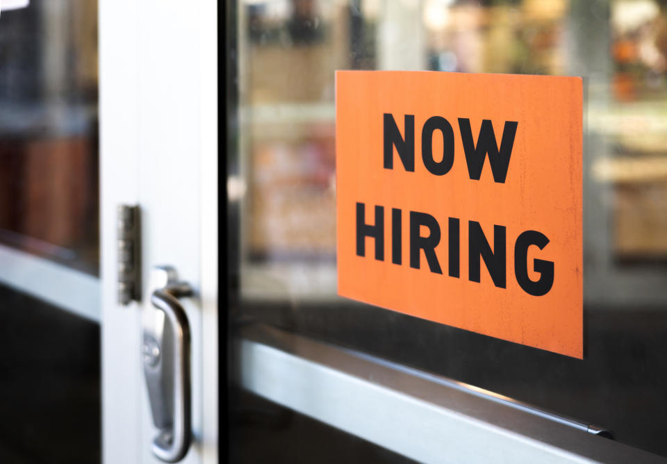 A "Now Hiring" sign is displayed in a window of a business establishment, indicating available job positions