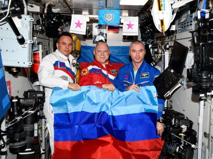 three cosmonauts pose with blue and red striped flag inside space station