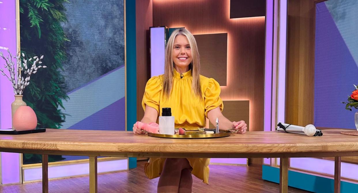 Claire Sisk wearing yellow presenting a makeup tutorial on This Morning while sitting at a wooden table. 