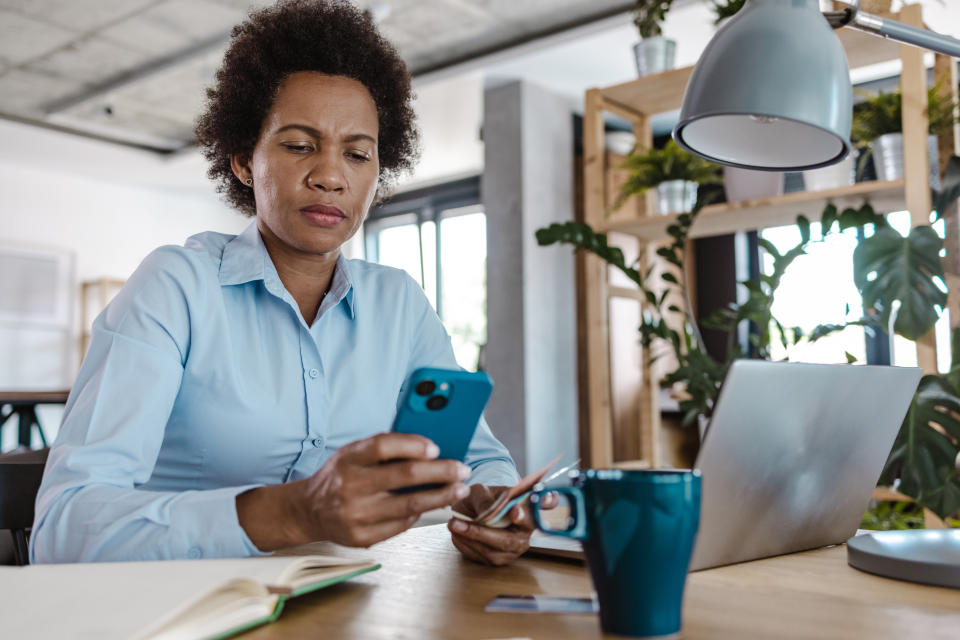 An African-American woman in a blue shirt sits at the table and works on a laptop. She is using smart phone