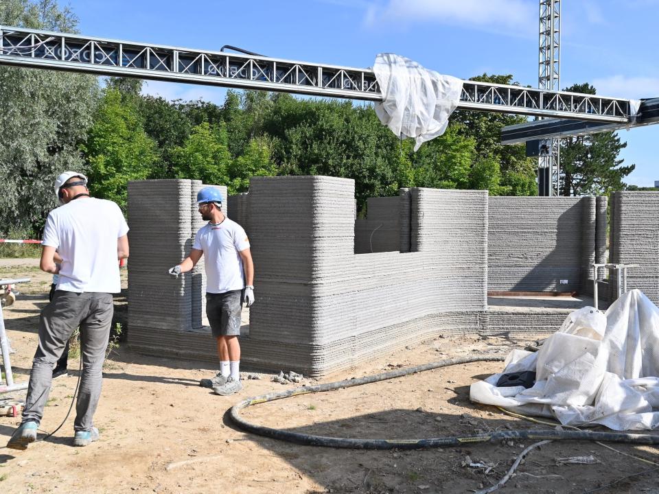 A 3D printer above the printed walls of the home. Two people are standing nearby.