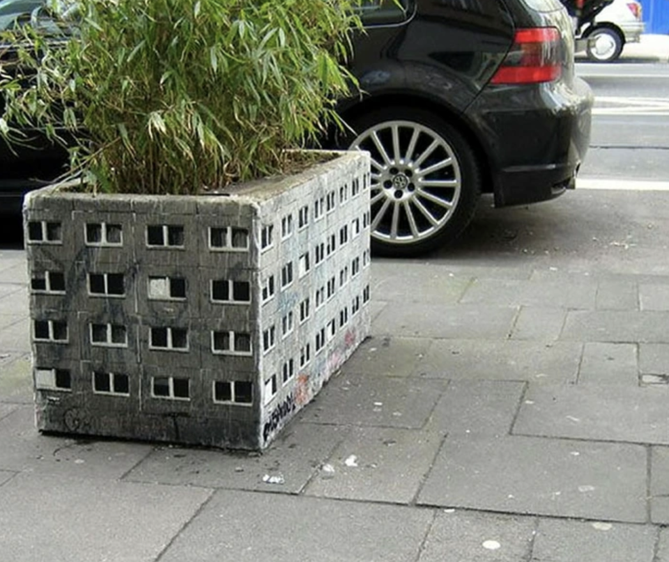 Planter made from cinder blocks resembles a city building, on a sidewalk with a car parked nearby