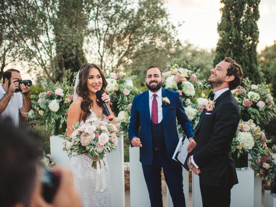 A bride speaks into a microphone as her groom laughs at her wedding.