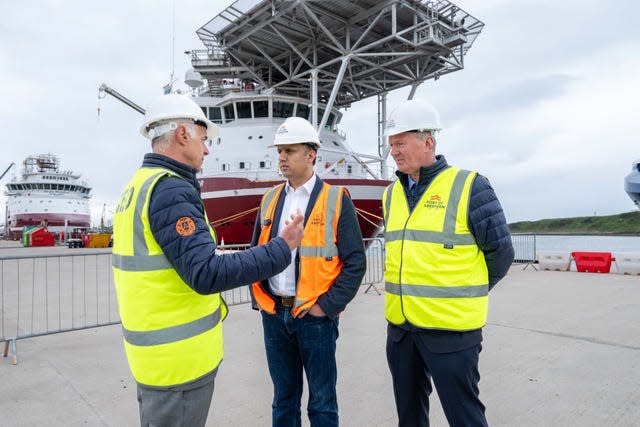 Scottish Labour leader Anas Sarwar in an orange work vest talks to two men in yellow high-vis in front of a large boat under a canopy