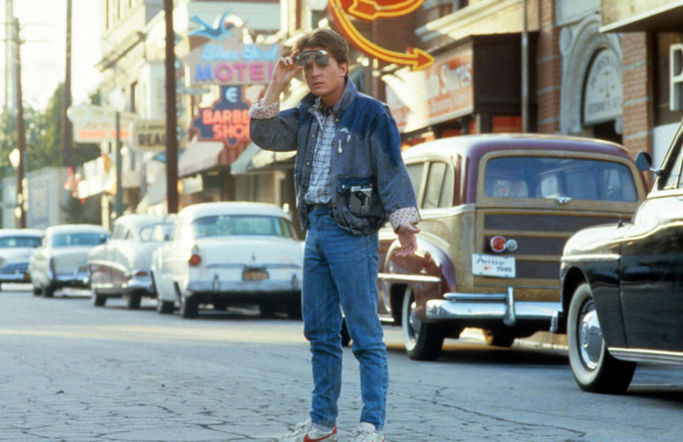 Marty McFly stands on a street in "Back to the Future", wearing a denim jacket, vest, and holding sunglasses
