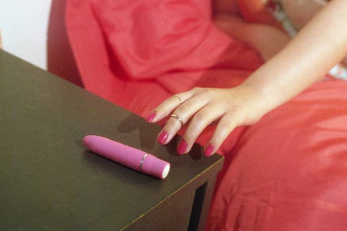 A person with manicured nails and rings reaches for a pink vibrator on a bedside table next to pink bedding