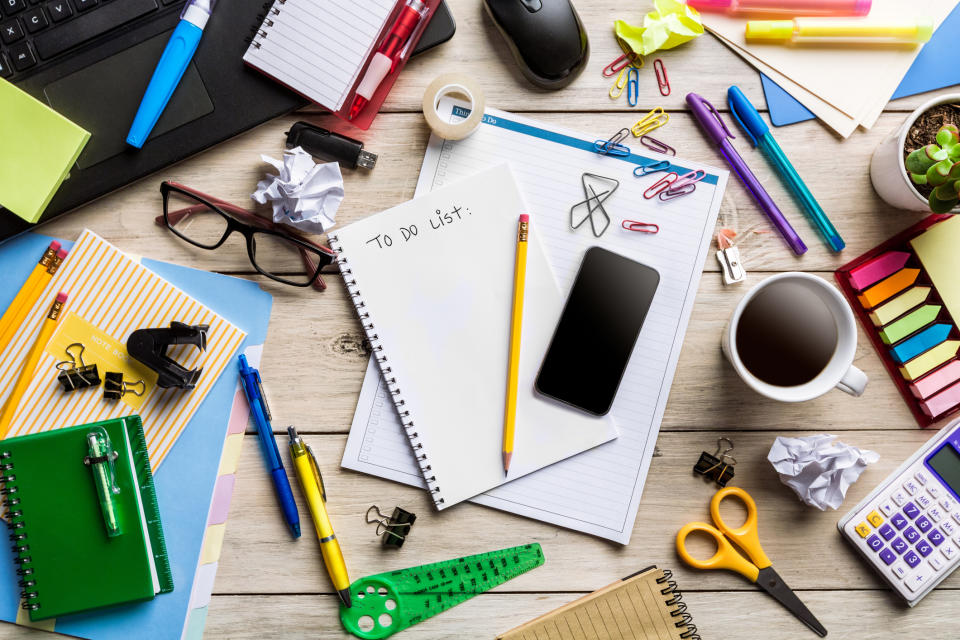 A cluttered desk with a to-do list, pens, glasses, phone, coffee cup, clips, sticky notes, notebooks, scissors, tape, a plant, and a laptop