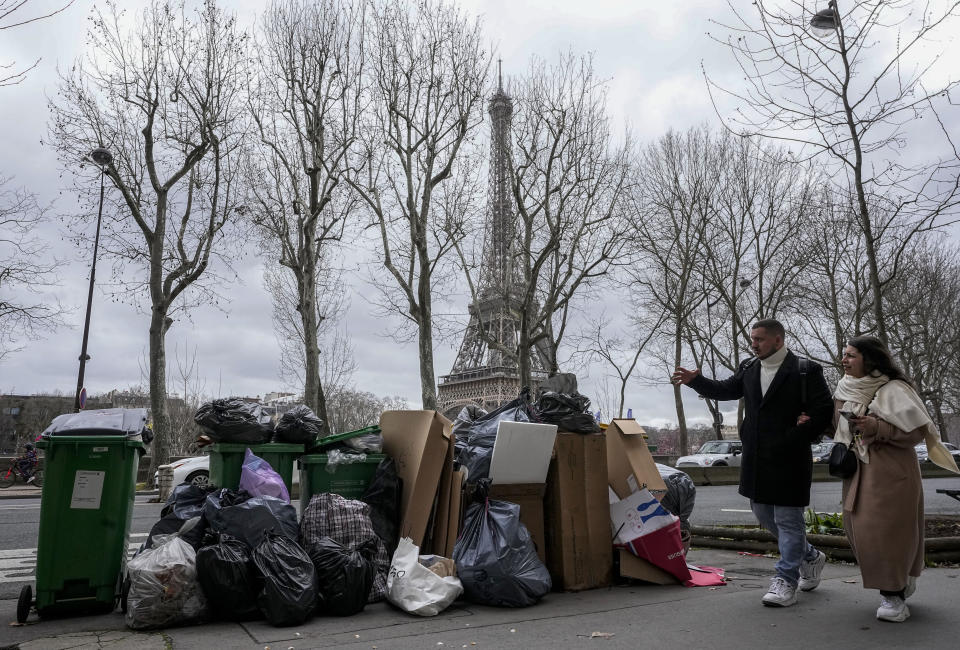 People walk past not collected garbage cans near the Eiffel Tower in Paris, Sunday, March 12, 2023. A contentious bill that would raise the retirement age in France from 62 to 64 got a push forward with the Senate's adoption of the measure amid strikes, protests and uncollected garbage piling higher by the day. (AP Photo/Michel Euler)