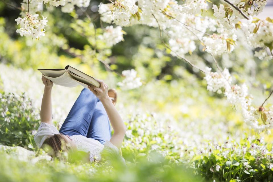 a woman wearing jeans and a white top lies in the grass under a flowering tree and reads a book, a summer bucket list idea