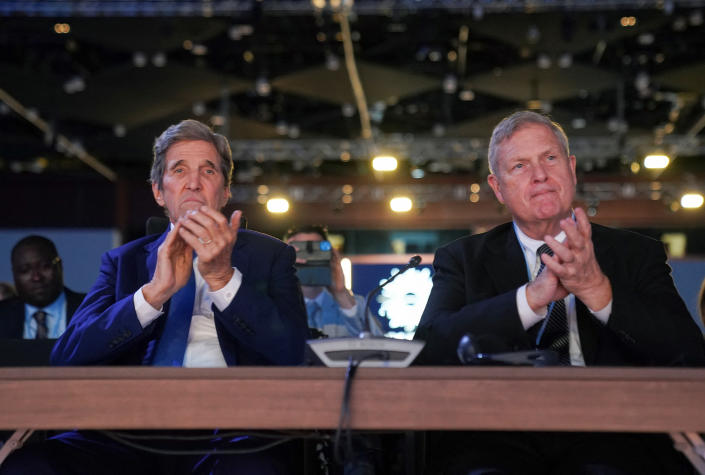 John Kerry and Secretary of Agriculture Tom Vilsack, seated at a single table in the auditorium, applaud.