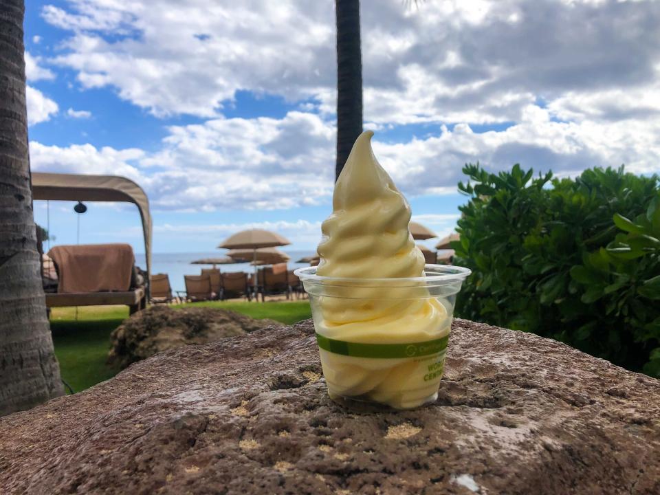 A cup of yellow soft serve dole whip on a rock with views of the ocean behind it.